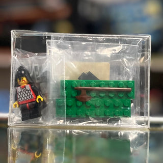LEGO Castle minifigure with red and black armor alongside green baseplate and brown catapult piece in clear plastic case.