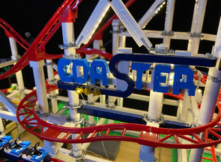 Illuminated blue "COASTER" sign on roller coaster model with red track, white support beams, and yellow lighting elements.