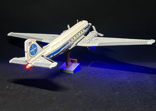 White and blue LEGO Douglas airliner with illuminated windows, landing gear, and red tail light on blue display stand.