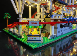 LEGO roller coaster set with illuminated ticket booth, colorful LED lights on structures, and lit minifigure on green basepla
