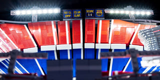 Illuminated LEGO stadium model with red and blue seating, scoreboard, and bright overhead lighting bars.